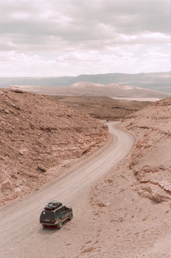 An SUV navigating a winding dirt road in a rugged desert landscape under a cloudy sky.