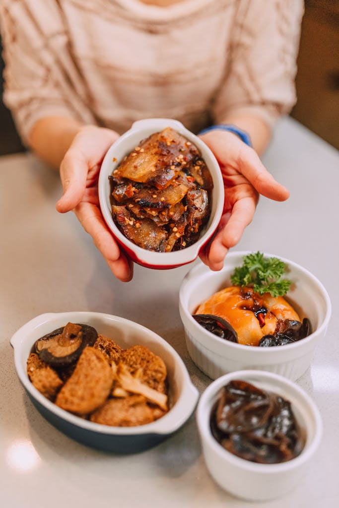 Hands holding and displaying a variety of appetizing Asian dishes in ceramic bowls on a table.
